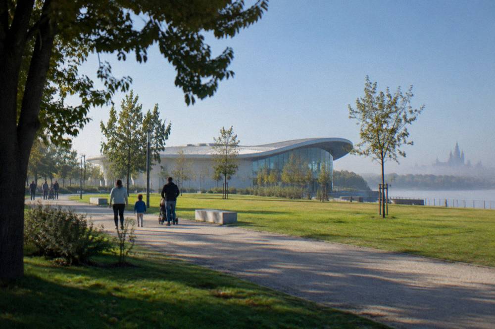 Families walking along a wide path through a large riverside park with lawns and trees