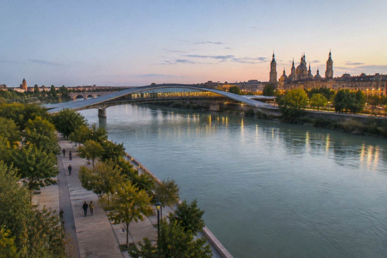 A curved modern bridge building crossing the Ebro river at dusk with the Zaragoza skyline behind it