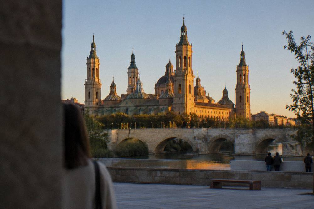 A stone bridge over a river with a large basilica tower rising behind it in golden evening light