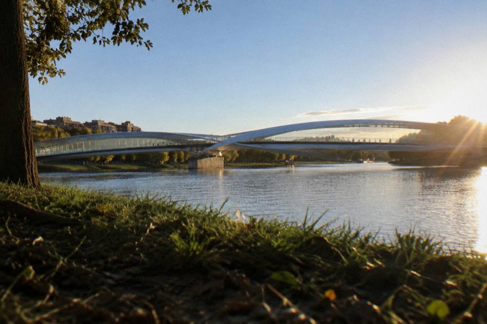 A modern covered bridge over a calm river seen from a park bank in late afternoon light
