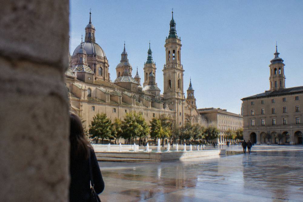 A quiet plaza in a historic Zaragoza district with ochre stone buildings and a church tower behind