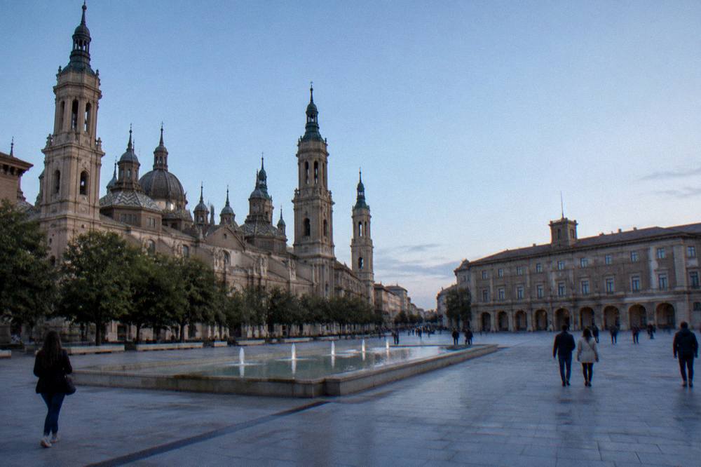 Visitors on a wide stone plaza in Zaragoza between historic buildings in late afternoon light
