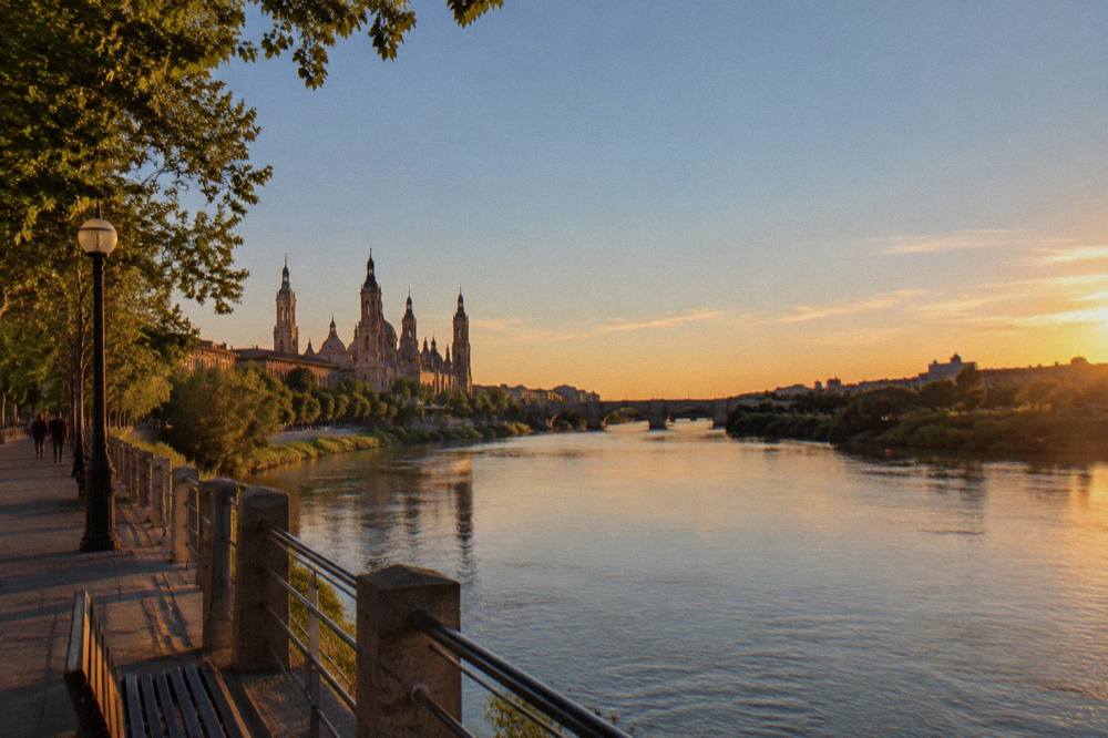 Golden evening light over the Ebro with parkland and a distant bridge silhouette