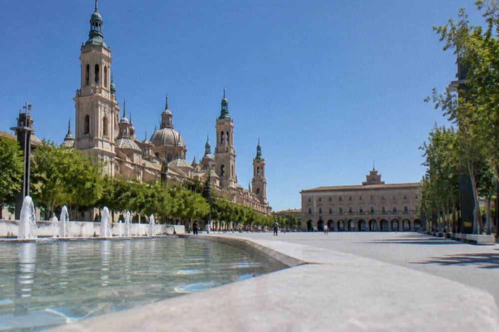 Wide formal promenade with tall trees and a stone fountain in a Zaragoza park