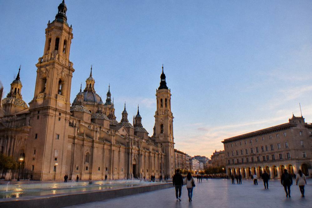 People strolling across the open square in front of the basilica at golden hour in Zaragoza