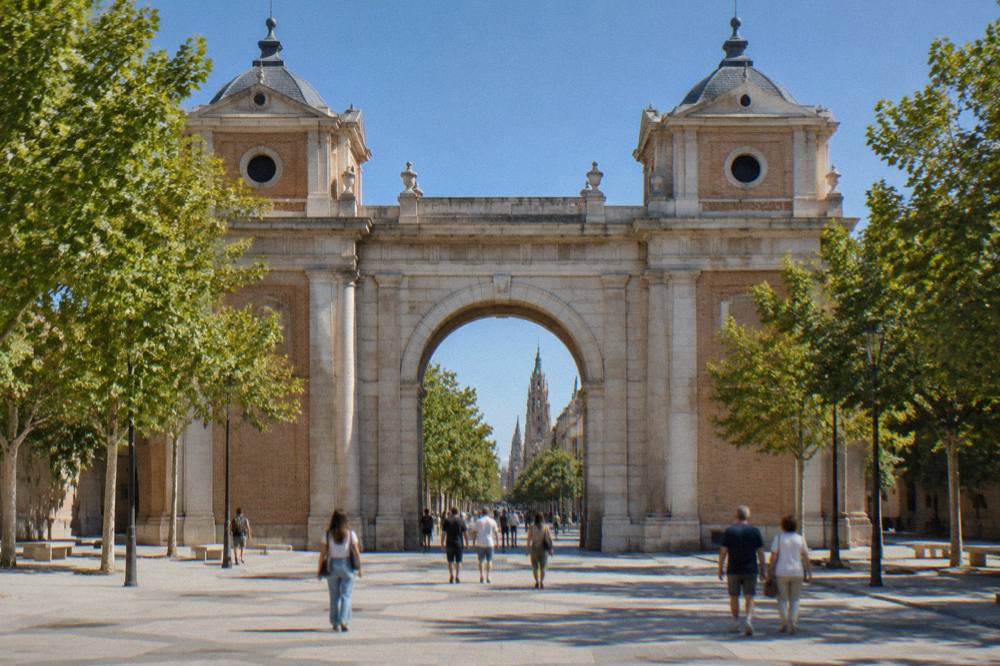 Pedestrians walking through a stone archway gate on a sunny afternoon in a Spanish city