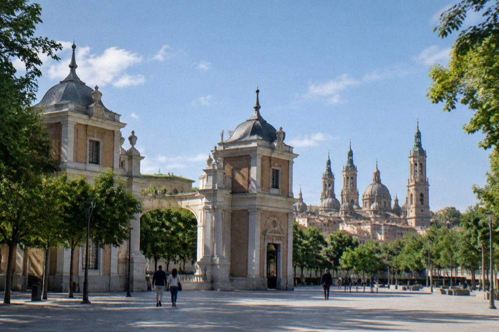 A wide city square with a large basilica and a cathedral tower seen across rooftops