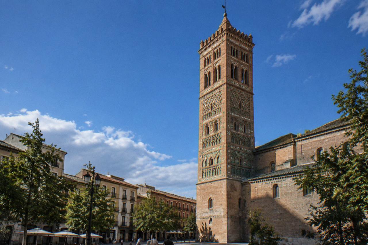 A tall brick church tower with intricate Mudéjar patterned decoration rising over a city square