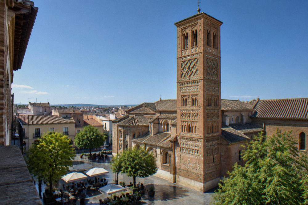 A quiet square with cafe tables in front of a brick Mudéjar church tower