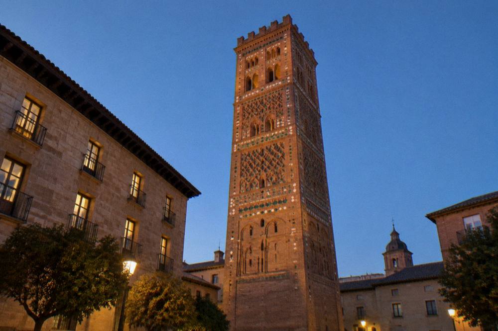 A Mudéjar brick tower photographed in warm evening light with long shadows across the square