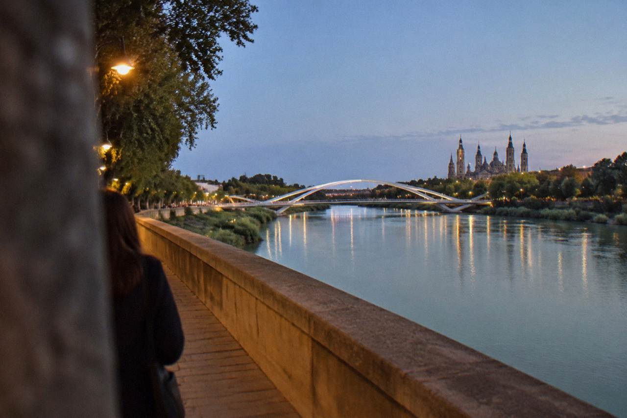 A sweeping modern bridge with curved white supports crossing the Ebro river at dusk, with parkland on both banks