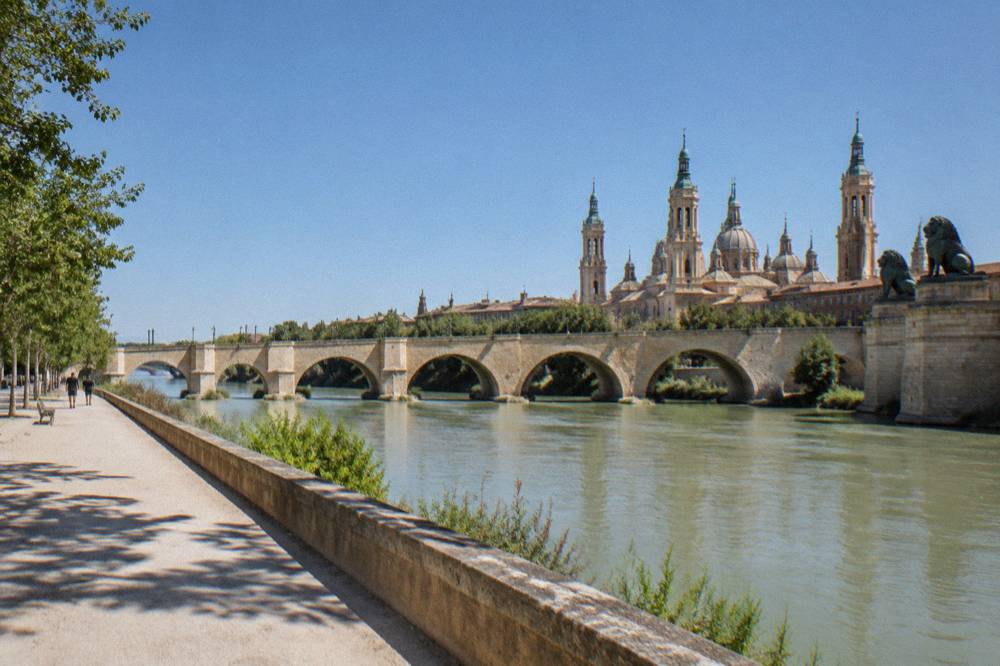 A wide riverside path along the Ebro with the stone arches of the Puente de Piedra bridge in the distance