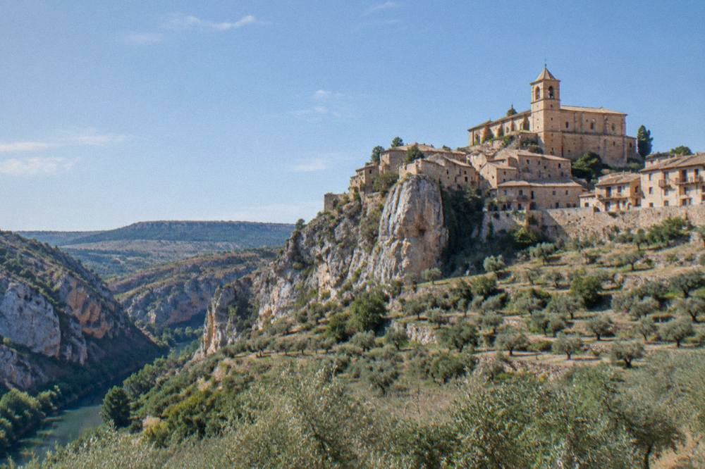 A hilltop village church with a bell tower seen across a valley of olive groves