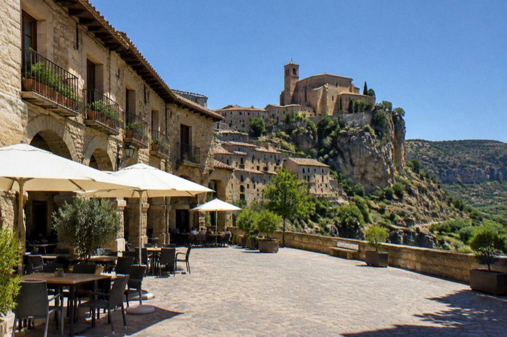 A village square with stone arcades and tables under shade at the foot of a rocky hill