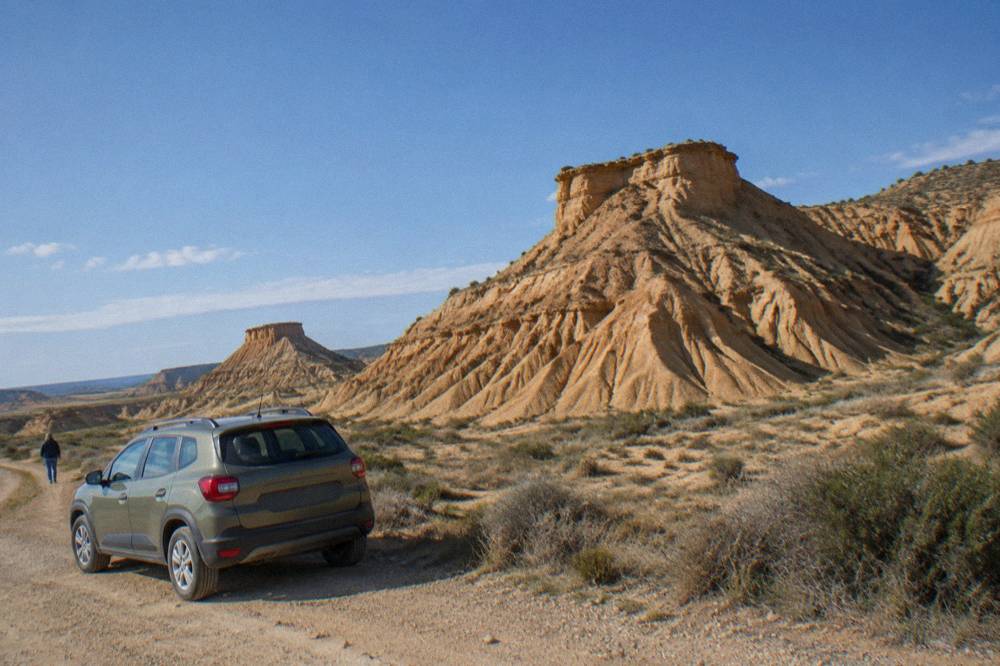 Parked car beside a dirt track with eroded ochre hills rising in the background under strong sunlight