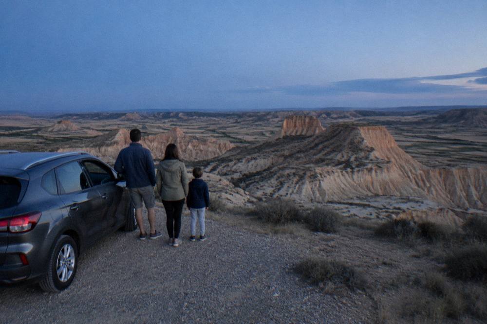 Family standing beside a car at a viewpoint looking out over pale eroded hills in the badlands