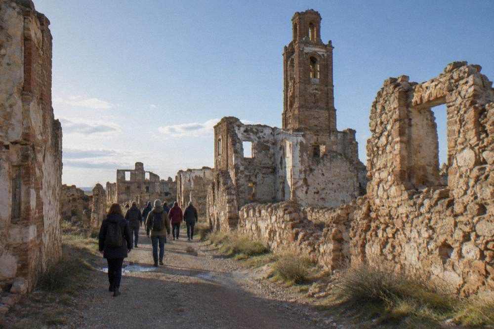 A guided group walking a dusty lane between ruined walls at Belchite old town