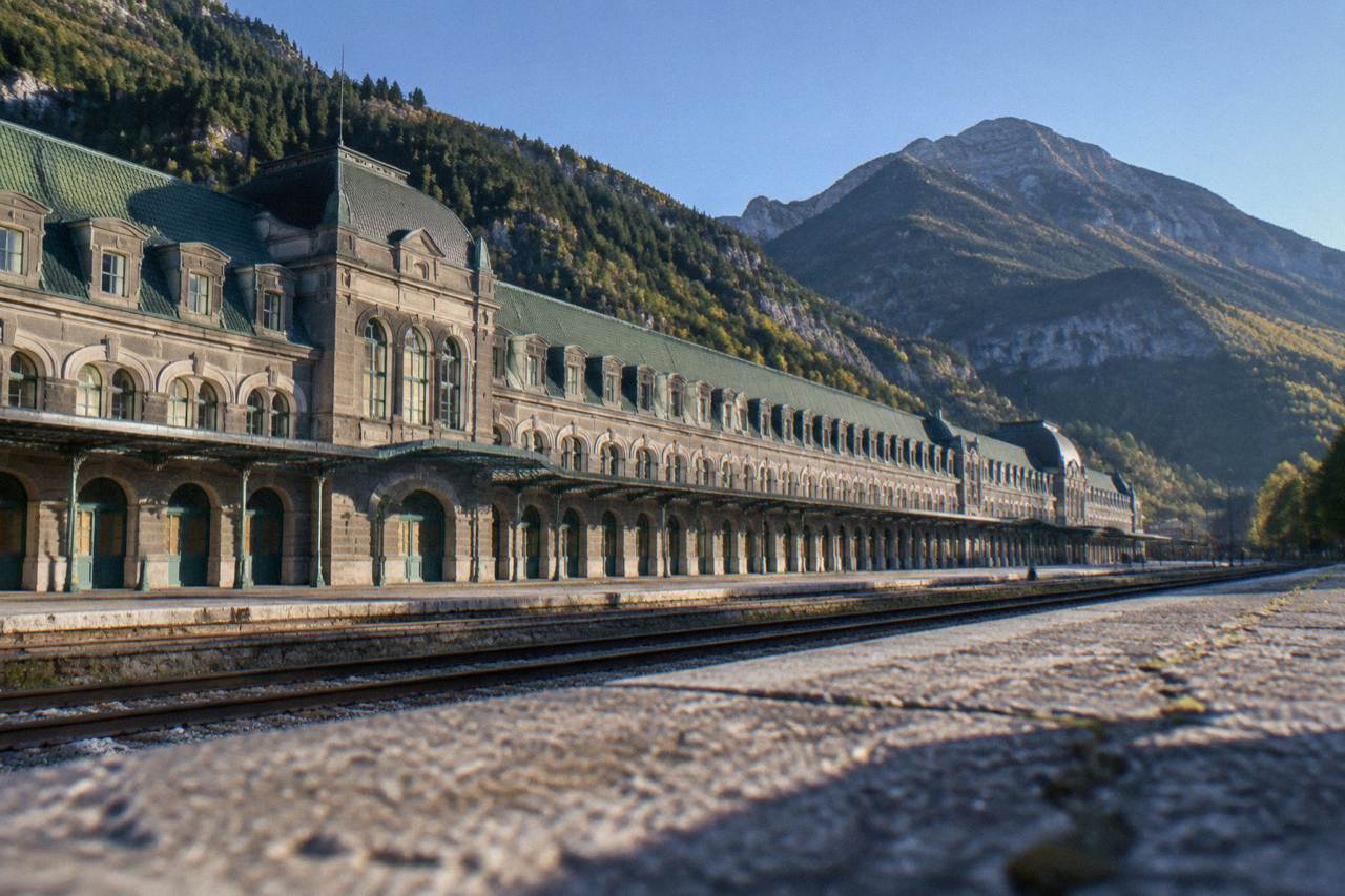 A long stone railway station building with a green tiled roof and rows of arched windows under mountains