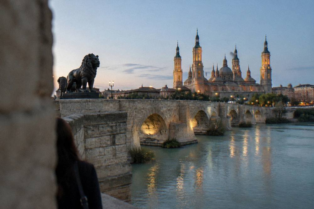 A stone bridge over a wide river with a lit basilica tower behind at dusk
