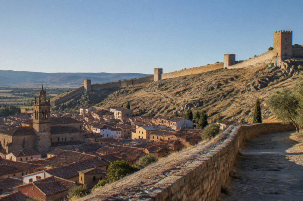 A long medieval stone wall curving along a hillside above red-roofed houses in a Spanish town