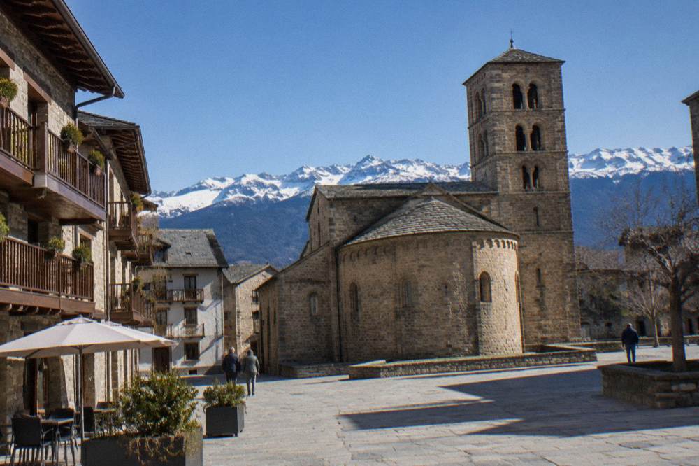 A stone village square with wooden balconies and mountains behind it in the Aragonese Pyrenees