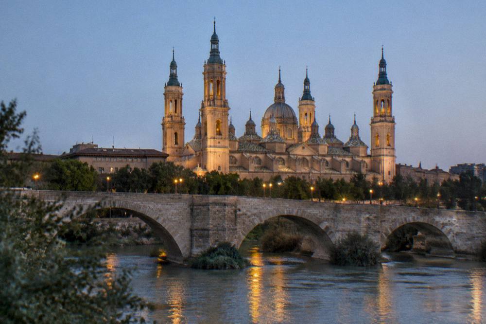 The stone arches of the Puente de Piedra in Zaragoza at dusk with the basilica towers lit behind
