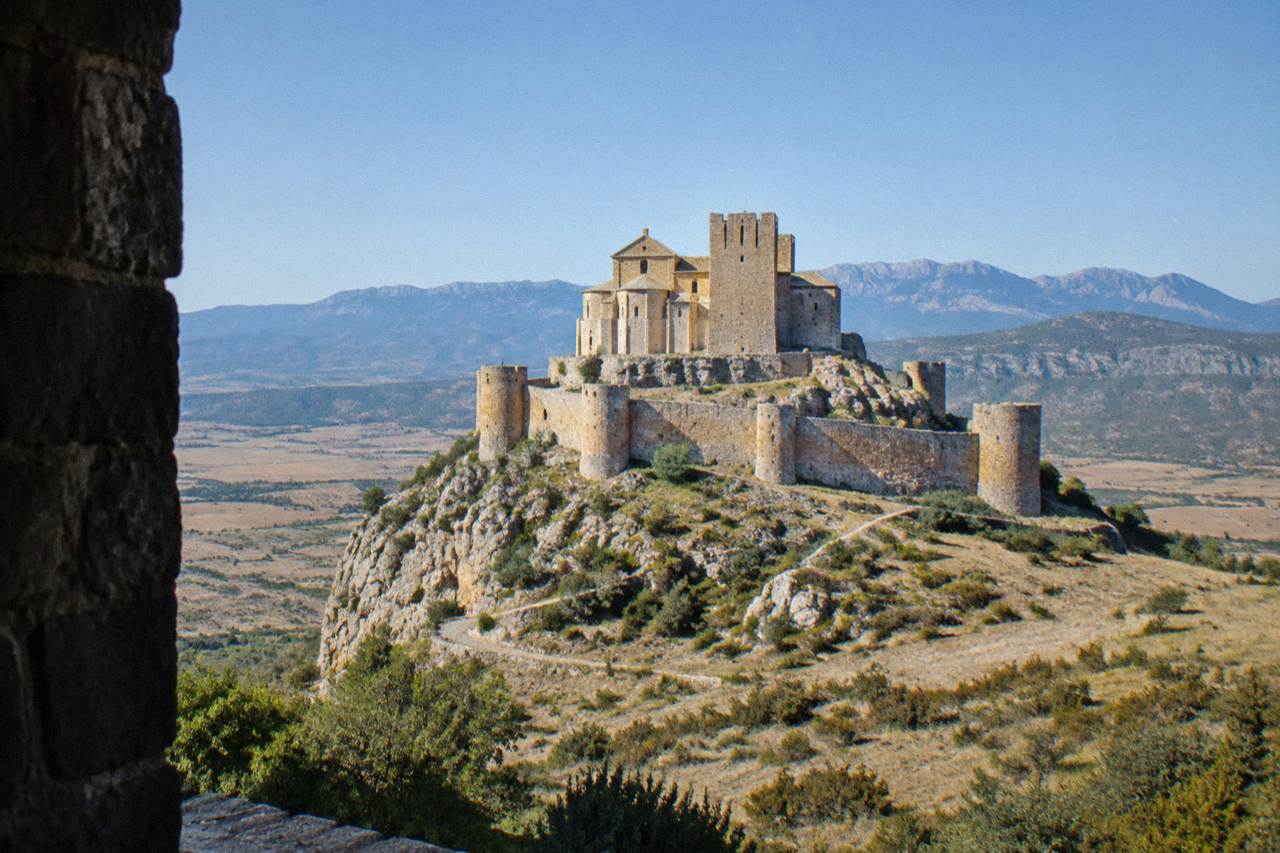 A stone Romanesque castle with round towers standing on a rocky ridge above dry hills
