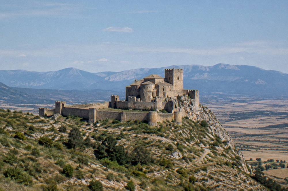 A wide view of a castle on a ridge with open country stretching to distant mountains