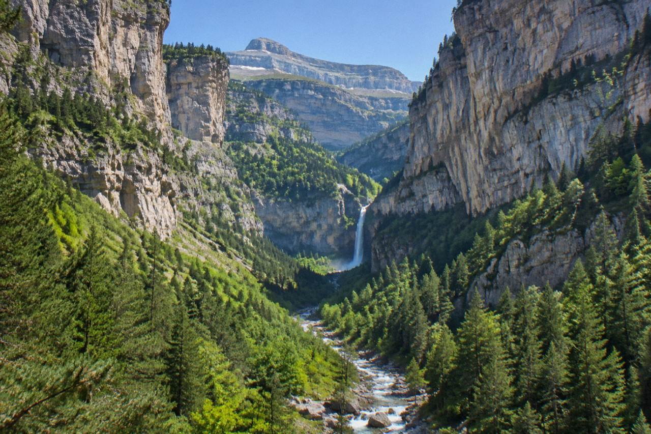 A steep green mountain valley with a river running through pine forest and high rock walls under a blue sky
