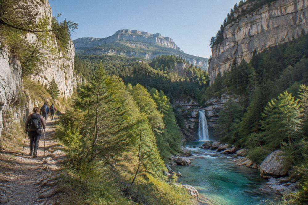 Hikers on a mountain path above a turquoise river with forested slopes rising behind them