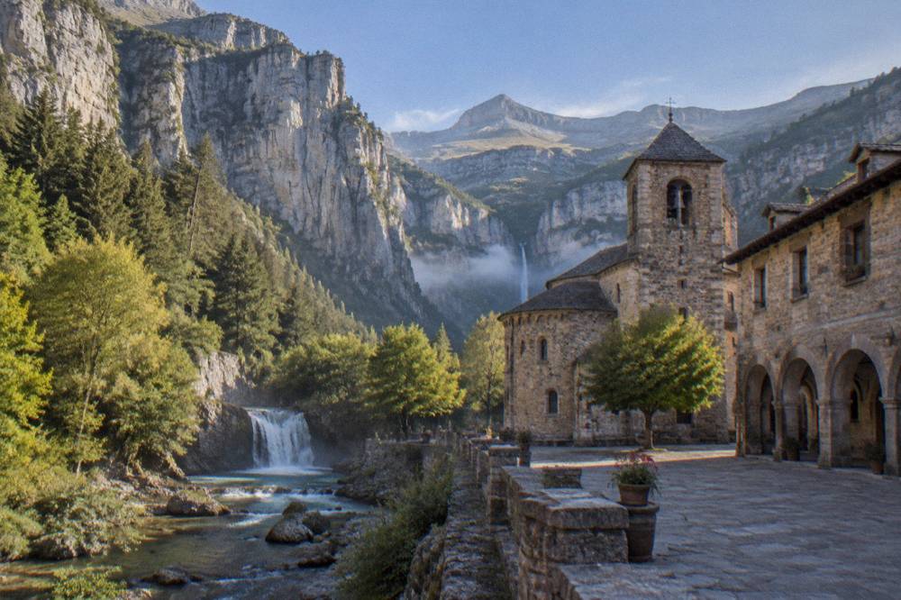 An old stone plaza with arcades and a church under mountains in late afternoon light