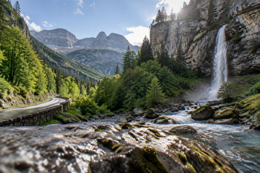 A narrow mountain road winding through forest toward high grey peaks