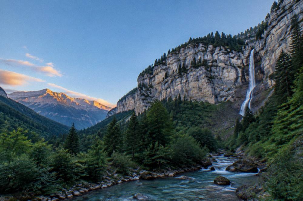 Golden evening light over a mountain valley with a river winding between dark forested slopes