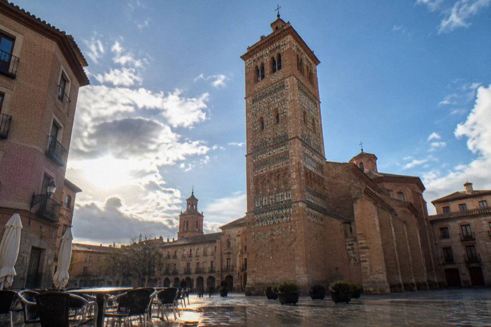 A brick bell tower with patterned ceramic decoration seen from a plaza with cafe tables