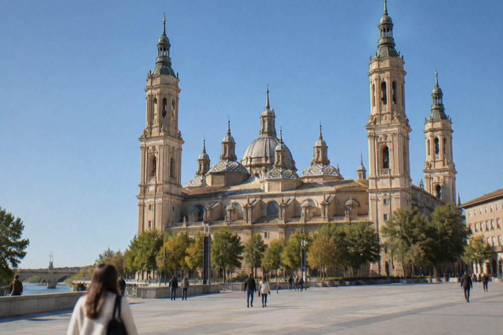 A grand baroque basilica with tiled domes rising over a wide stone square