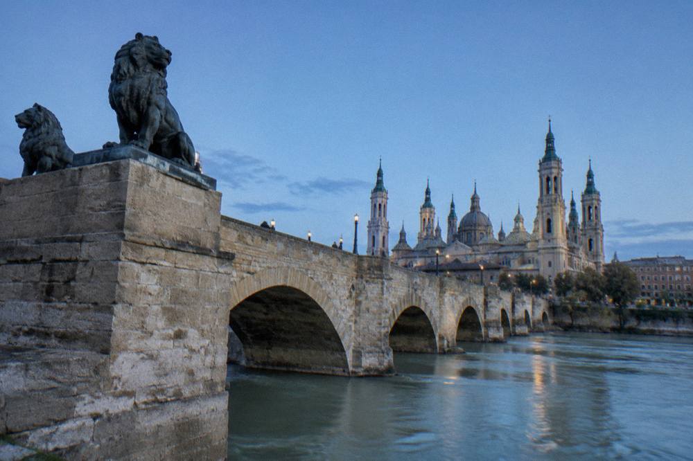 A wide river flowing under a historic stone bridge with a city skyline of domes and towers behind