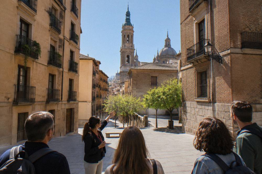 A guide speaking to a small group on a plaza with a cathedral tower rising behind the rooftops