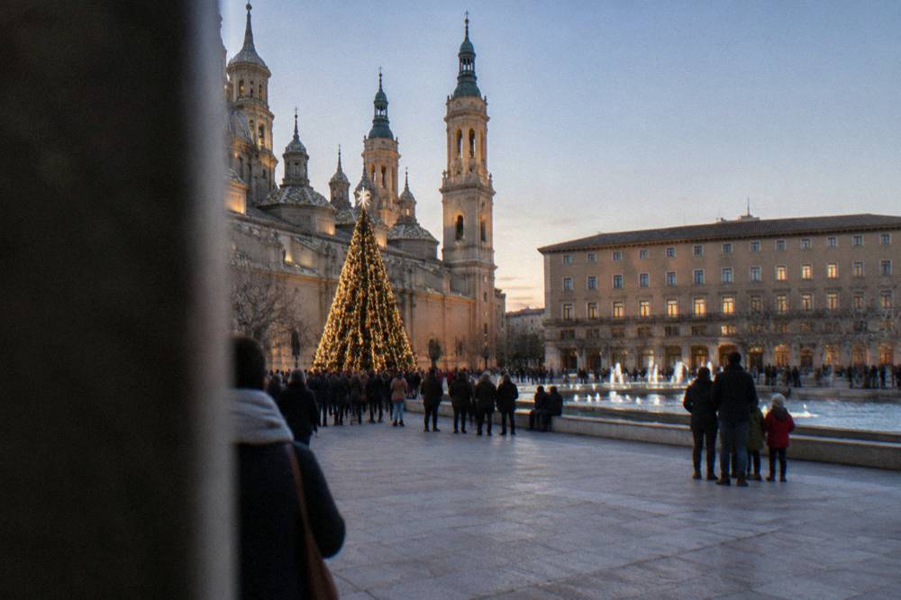 A lit Christmas tree in a Zaragoza square with people in winter coats gathered around it in the evening