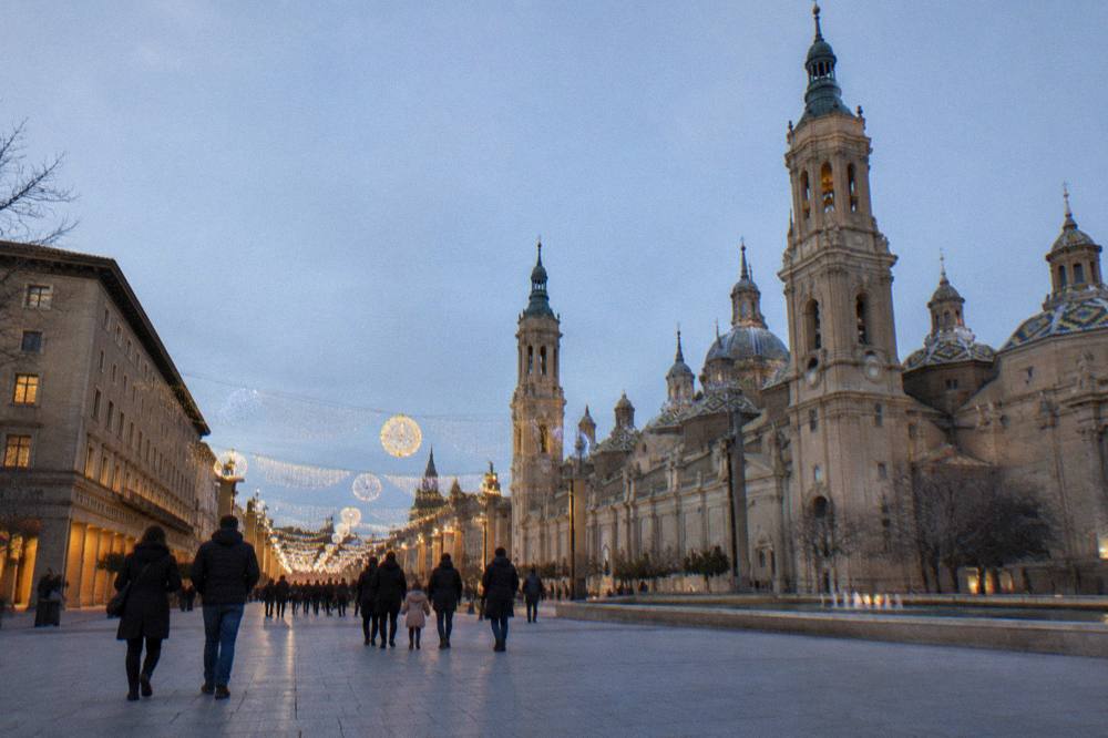 Families walking under illuminated Christmas decorations spanning a shopping street in Zaragoza at night