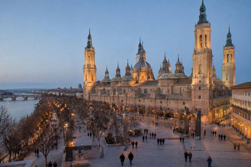 A wide evening view of Plaza del Pilar in winter with the basilica towers lit and the square softly illuminated