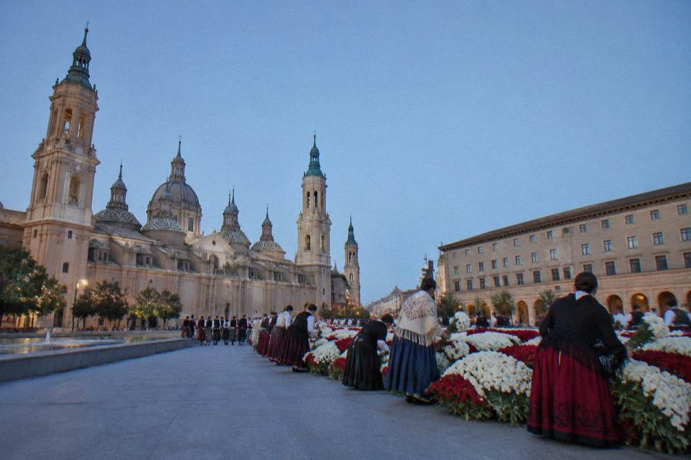 Rows of participants in traditional dress laying white and red flowers into a large structure in a square
