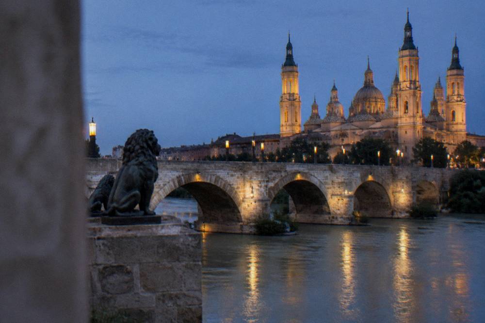 A stone bridge over a river at dusk with a floodlit basilica and its domes behind it