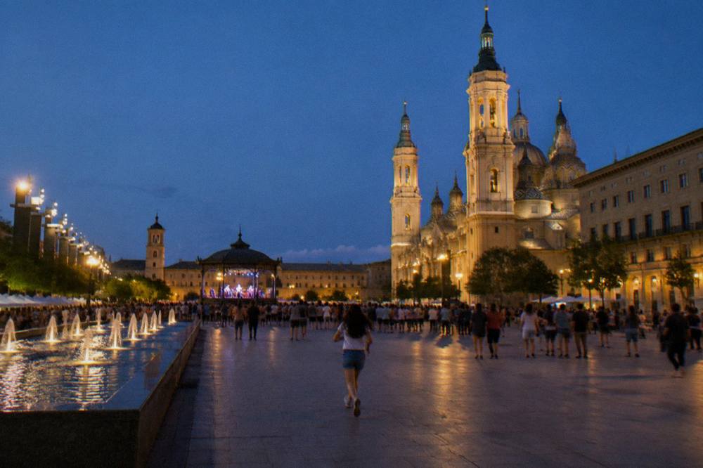 A lit festival square at night with crowds, a bandstand and historic buildings around the edge