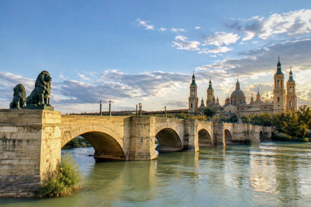 A stone bridge with bronze lion statues spanning the Ebro river at golden hour