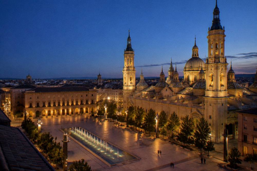 An evening view of Zaragoza's old town rooftops with the basilica dome lit against a dark blue sky