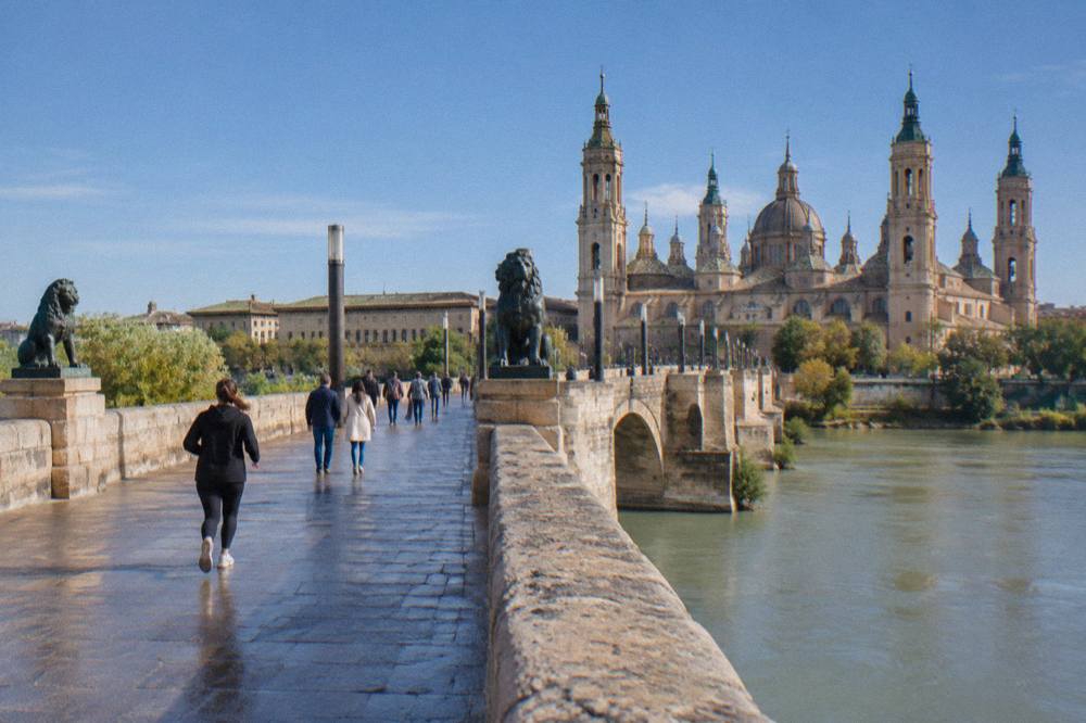 People strolling across the Puente de Piedra stone bridge with the Basilica del Pilar behind them