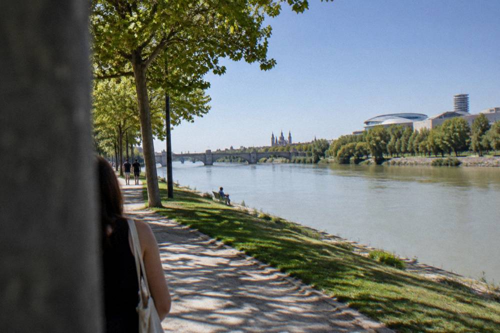 A wide green park path beside the Ebro river with trees and modern Expo buildings in the distance