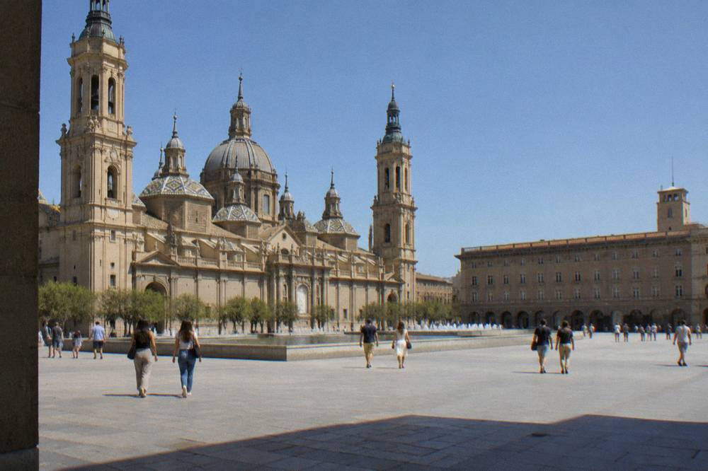 Pedestrians crossing a wide stone plaza in front of a large baroque basilica in Zaragoza
