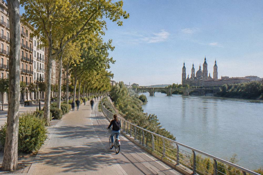A cyclist riding a shared bike along a riverside path beside a wide river in the late afternoon