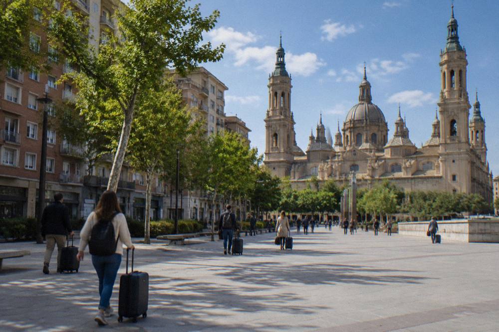 Travellers with suitcases walking through a sunlit historic square with a large baroque basilica in the background